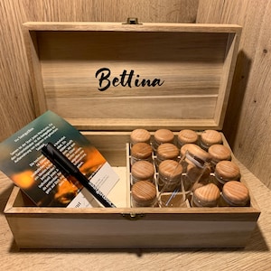 Op de afbeelding: Een houten kruidenkist met een scharnierend deksel, met kleine glazen potjes met houten deksels. De doos is open en onthult de potjes in rijen. Een brochure en een zwarte pen bevinden zich in de doos. De naam "Bettina" staat op de binnenkant van het deksel.