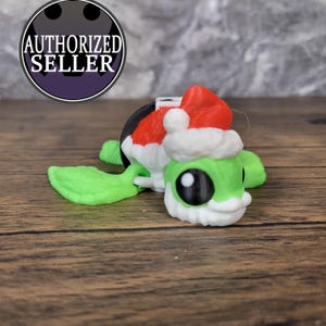 May include: A green and white toy turtle wearing a red and white Santa hat. The turtle has a black base and is made of plastic.