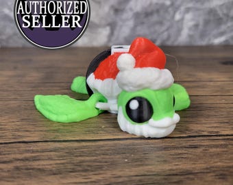 May include: A green and white toy turtle wearing a red and white Santa hat. The turtle has a black base and is made of plastic.