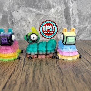 May include: Three colorful llama figurines with rainbow-colored skirts and geometric faces. The llamas are in shades of blue, green, and yellow, with black accents. The Flexi Factory logo is visible in the background.