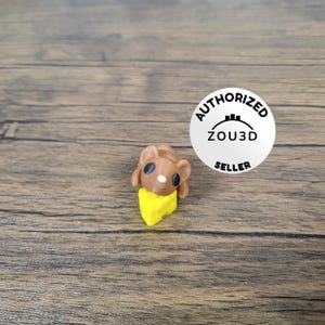 May include: A brown mouse figurine holding a yellow cheese wedge. The figurine is on a wooden surface. The image also includes a white sticker with the text "AUTHORIZED ZOU3D SELLER".