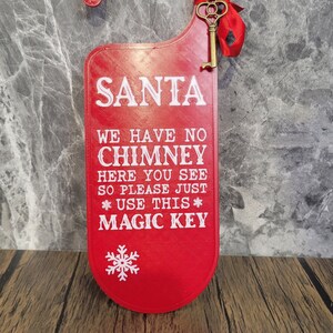 May include: Red door hanger with a gold key and a white snowflake. The text on the door hanger reads "SANTA WE HAVE NO CHIMNEY HERE YOU SEE SO PLEASE JUST * USE THIS * MAGIC KEY".