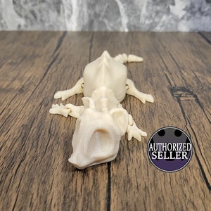 May include: A white plastic articulated skeleton of a lizard with a long tail and open mouth. The skeleton is posed on a wooden surface.