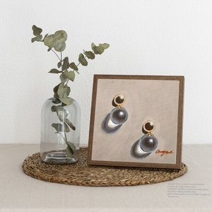 May include: A framed painting of a pair of pearl earrings with gold accents. The earrings are on a beige background with a shadow cast below each earring. The painting is in a wooden frame.