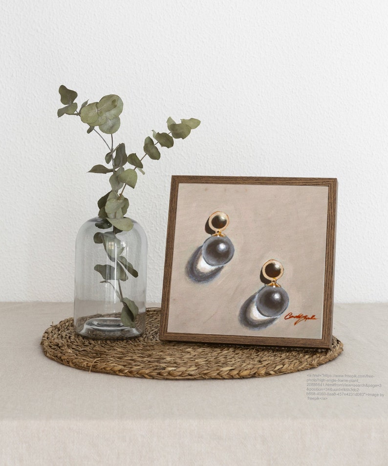 May include: A framed painting of a pair of pearl earrings with gold accents. The earrings are on a beige background with a shadow cast below each earring. The painting is in a wooden frame.