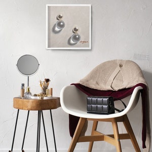 May include: A white chair with a beige sweater draped over the back. A black purse sits on the seat of the chair. A small wooden table with a round mirror and a few items on top sits next to the chair. A framed drawing of a pair of pearl earrings hangs on the wall behind the chair.