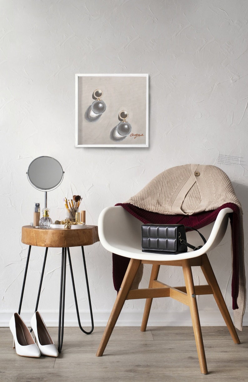 May include: A white chair with a beige sweater draped over the back. A black purse sits on the seat of the chair. A small wooden table with a round mirror and a few items on top sits next to the chair. A framed drawing of a pair of pearl earrings hangs on the wall behind the chair.
