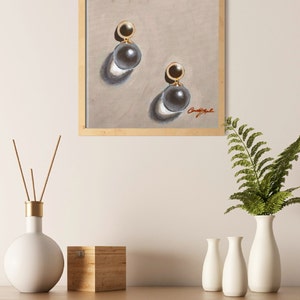 May include: A framed painting of a pair of pearl earrings with gold accents. The earrings are depicted on a light beige background. The painting is signed "CandyApple".