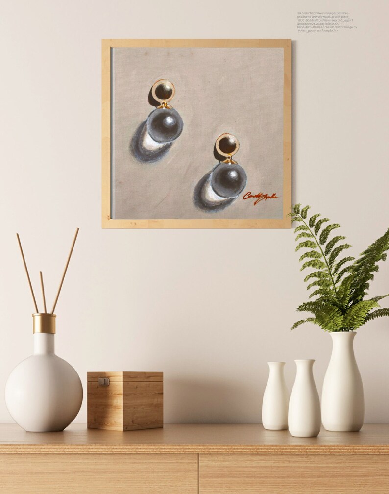 May include: A framed painting of a pair of pearl earrings with gold accents. The earrings are depicted on a light beige background. The painting is signed "CandyApple".