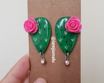 Peut inclure: Une paire de boucles d'oreilles en forme de cactus vert avec des accents de fleurs roses et un pendentif en strass transparent. Les boucles d'oreilles sont sur une carte marron avec le texte "@iamjomalulu".