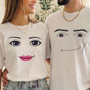 May include: Two white short-sleeve t-shirts. The left shirt features a graphic of a face with dark eyebrows, large eyes, and bright pink lips. The right shirt features a graphic of a face with dark eyebrows, large eyes, and a thin black line smile.
