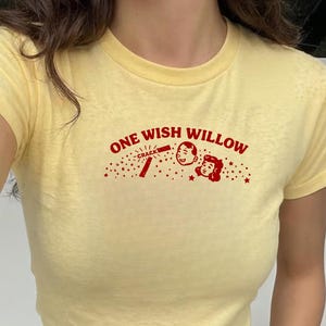 May include: A pale yellow short-sleeved t-shirt featuring a red graphic on the chest. The graphic includes the text "ONE WISH WILLOW" above a small illustration of two faces, a "CRACK" label, and scattered stars. The shirt has a crew neckline.
