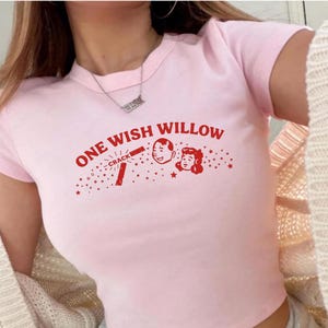Op de afbeelding: Een lichtroze crop top met korte mouwen en een rode grafische print met de tekst ONE WISH WILLOW, een kleine illustratie van een gezicht en een stokje met het label CRACK, omringd door kleine sterretjes, gedragen met een grijze joggingbroek.