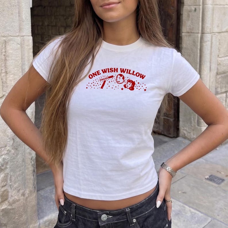 Op de afbeelding: Een wit T-shirt met korte mouwen en een rode grafische print op de borst. Het ontwerp bevat de tekst "ONE WISH WILLOW" in een gebogen lettertype, vergezeld van een kleine illustratie van twee gezichten en het woord "CRACK" omringd door sterren.