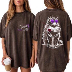 May include: A dark brown mineral-washed t-shirt featuring a small front graphic that reads The Desperado Club. The back displays a large white line-art illustration of a fluffy cat wearing a crown and purple sunglasses, sitting beside a martini glass.