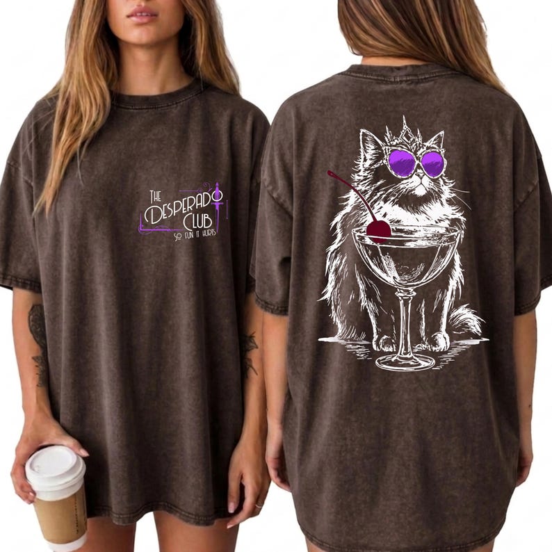 May include: A dark brown mineral-washed t-shirt featuring a small front graphic that reads The Desperado Club. The back displays a large white line-art illustration of a fluffy cat wearing a crown and purple sunglasses, sitting beside a martini glass.