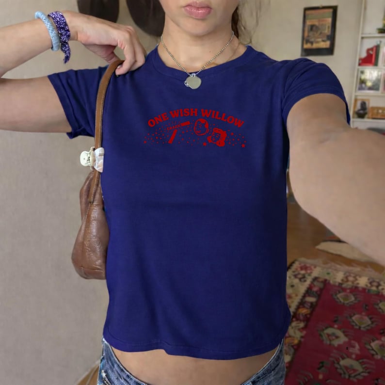 Op de afbeelding: Een donkerblauw T-shirt met korte mouwen en de rode tekst ONE WISH WILLOW op de borst, vergezeld van kleine rode grafische iconen van een toverstaf, een ster en een klein rechthoekig kader omringd door verspreide rode stippen.