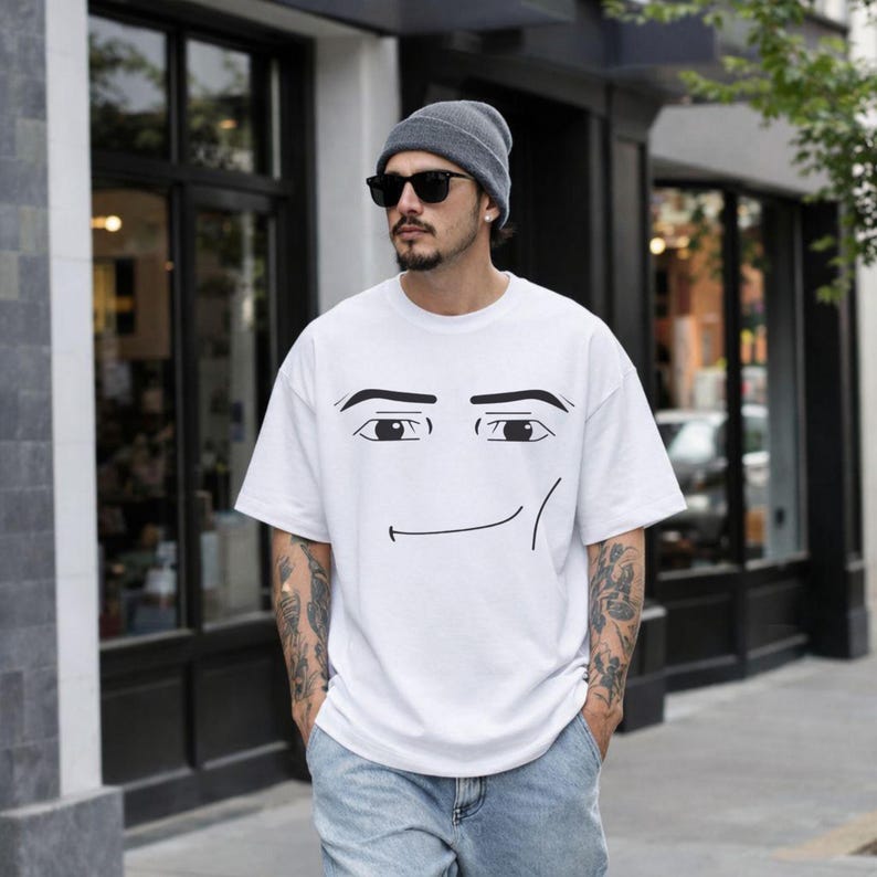 May include: A white short-sleeved t-shirt featuring a black graphic print of a stylised face with thick eyebrows, wide eyes, and a smirk. The shirt has a crew neckline and a loose, relaxed fit, constructed from a plain white fabric.