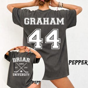 May include: A charcoal gray short-sleeve t-shirt featuring white screen-printed text and graphics. The back displays the name GRAHAM and the number 44. The front shows the text BRIAR UNIVERSITY, HAWKS, EST. 2015, and two crossed hockey sticks.