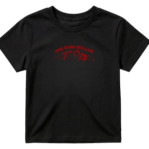 Op de afbeelding: Een zwart t-shirt met korte mouwen en een rode grafische print op de borst. Het ontwerp bevat de tekst ONE WISH WILLOW in een boog boven een kleine illustratie van een rechthoekig object en een cirkelvorm met verspreide rode stippen.