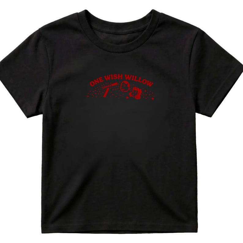 Op de afbeelding: Een zwart t-shirt met korte mouwen en een rode grafische print op de borst. Het ontwerp bevat de tekst ONE WISH WILLOW in een boog boven een kleine illustratie van een rechthoekig object en een cirkelvorm met verspreide rode stippen.