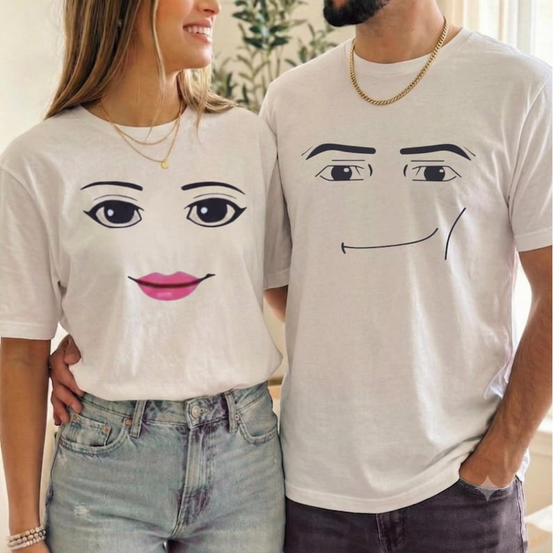 May include: Two white short-sleeve t-shirts. The left shirt features a graphic of a face with dark eyebrows, large eyes, and bright pink lips. The right shirt features a graphic of a face with dark eyebrows, large eyes, and a thin black line smile.