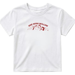 Op de afbeelding: Een wit t-shirt met korte mouwen en een rode grafische print op de borst. Het ontwerp bevat de tekst ONE WISH WILLOW, een kleine illustratie van een brekend wensbotje met het woord CRACK, en twee lachende gezichten omringd door sterretjes.