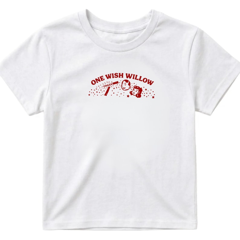 Op de afbeelding: Een wit t-shirt met korte mouwen en een rode grafische print op de borst. Het ontwerp bevat de tekst ONE WISH WILLOW, een kleine illustratie van een brekend wensbotje met het woord CRACK, en twee lachende gezichten omringd door sterretjes.