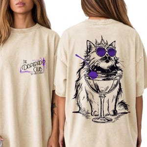 May include: A beige t-shirt featuring a small graphic on the front with the text The Desperado Club So Fun It Hurts. The back displays a large black and white illustration of a cat wearing a crown and purple sunglasses, sitting behind a martini glass.