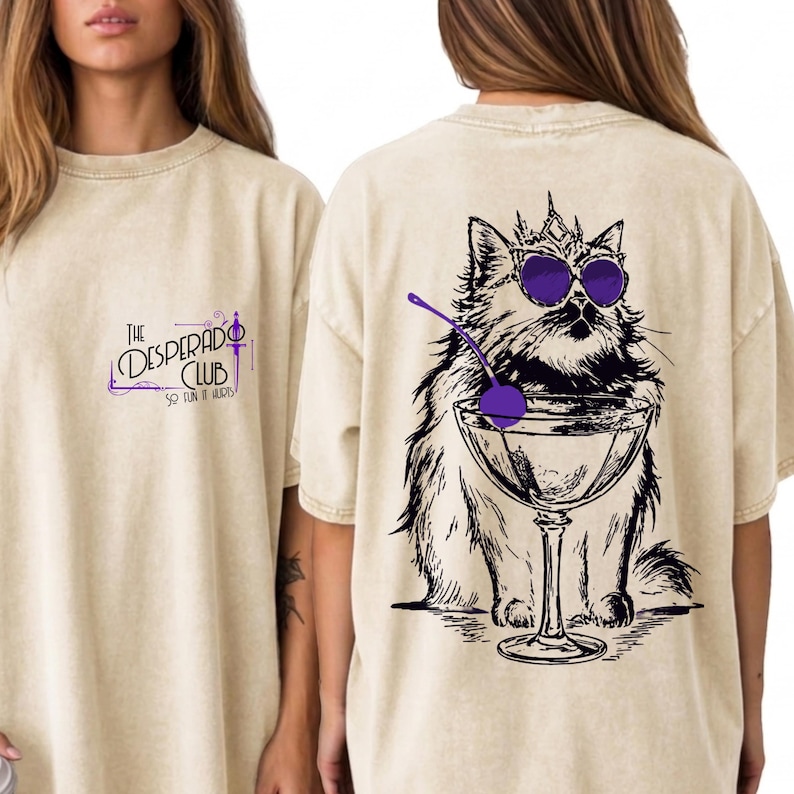 May include: A beige t-shirt featuring a small graphic on the front with the text The Desperado Club So Fun It Hurts. The back displays a large black and white illustration of a cat wearing a crown and purple sunglasses, sitting behind a martini glass.