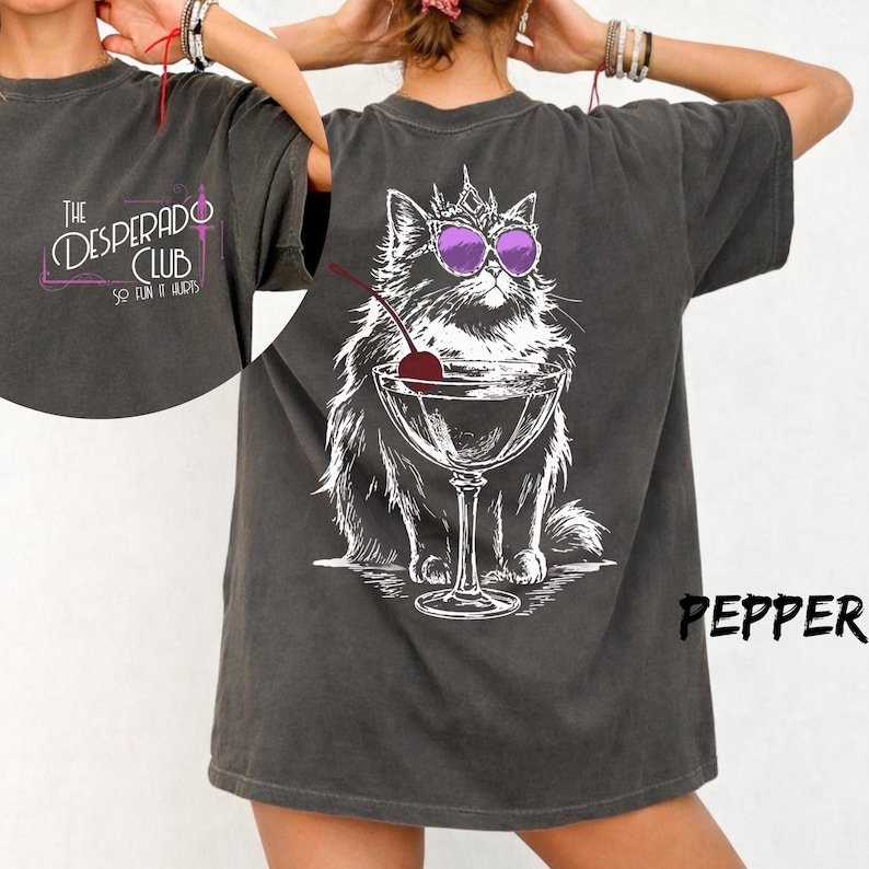 May include: A charcoal gray short-sleeve t-shirt featuring a white graphic of a cat wearing a crown and purple sunglasses next to a martini glass with a cherry. The front chest displays the text The Desperado Club So Fun It Hurts.