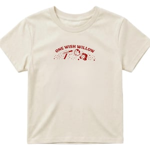 Op de afbeelding: Een cr&egrave;mekleurig T-shirt met korte mouwen en een rode afbeelding op de borst. De afbeelding bevat de tekst "ONE WISH WILLOW" in een boog boven een kleine illustratie van twee gezichten, een stokje met "CRACK" en verspreide sterren.