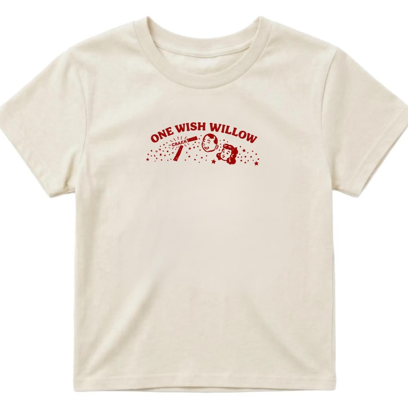 Op de afbeelding: Een cr&egrave;mekleurig T-shirt met korte mouwen en een rode afbeelding op de borst. De afbeelding bevat de tekst "ONE WISH WILLOW" in een boog boven een kleine illustratie van twee gezichten, een stokje met "CRACK" en verspreide sterren.