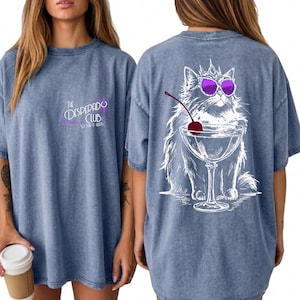 May include: A blue acid-wash t-shirt featuring a small logo on the front that reads The Desperado Club. The back displays a large white line-art illustration of a fluffy cat wearing a crown and purple sunglasses, sitting behind a martini glass.