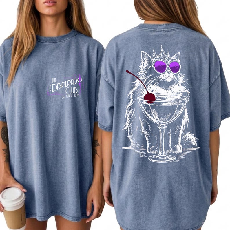 May include: A blue acid-wash t-shirt featuring a small logo on the front that reads The Desperado Club. The back displays a large white line-art illustration of a fluffy cat wearing a crown and purple sunglasses, sitting behind a martini glass.