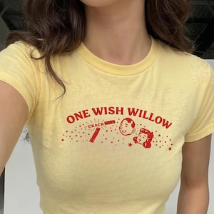 Op de afbeelding: Een lichtgeel t-shirt met korte mouwen en een rode grafische print op de borst. Het ontwerp bevat de tekst ONE WISH WILLOW, het woord CRACK, een illustratie van een gebroken stok en de gezichten van twee personen omringd door kleine sterren.