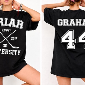 May include: A black t-shirt featuring a white graphic on the front with the text BRIAR HAWKS UNIVERSITY EST. 2015 and crossed hockey sticks with a puck. The back of the t-shirt displays the name GRAHAM and the number 44 in large white lettering.