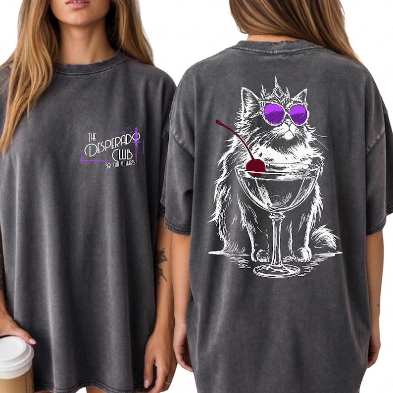 May include: A charcoal gray t-shirt featuring a small logo on the front that reads The Desperado Club So Fun It Hurts. The back displays a large white line art illustration of a fluffy cat wearing a crown and purple sunglasses behind a martini glass.