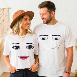 May include: Two white short-sleeved t-shirts featuring black graphic facial features. One shirt displays eyes and pink lips, while the other shirt displays eyes and a simple black line mouth. Both shirts are made of a plain white fabric material.