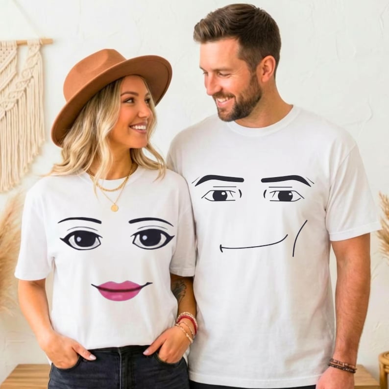 May include: Two white short-sleeved t-shirts featuring black graphic facial features. One shirt displays eyes and pink lips, while the other shirt displays eyes and a simple black line mouth. Both shirts are made of a plain white fabric material.