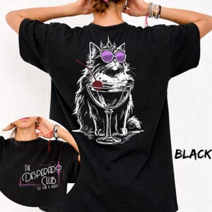 May include: A black t-shirt featuring a white line art illustration of a cat wearing a crown and purple sunglasses, holding a martini glass with a red cherry. The front chest area displays the text The Desperado Club So Fun It Hurts.