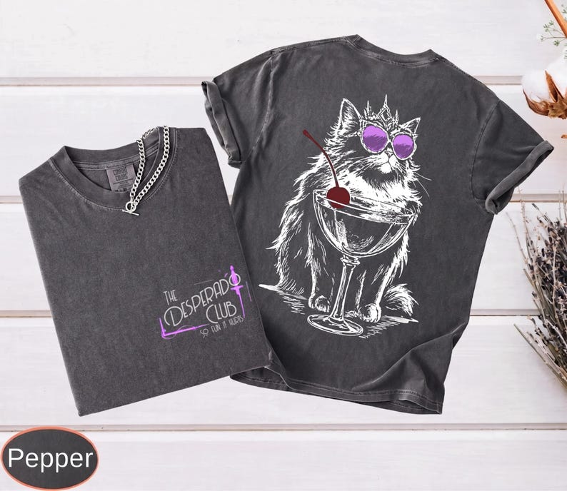 May include: A charcoal gray t-shirt featuring a graphic of a cat wearing a crown and purple sunglasses, sitting next to a martini glass with a cherry. The front displays the text The Desperado Club So Fun It Hurts in purple and white lettering.