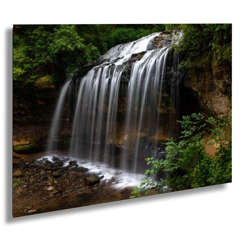 May include: A cascading waterfall flows over a rocky cliff, surrounded by lush green foliage. The water is white and foamy, creating a sense of movement and energy. The scene is tranquil and serene, with the natural beauty of the waterfall on display.