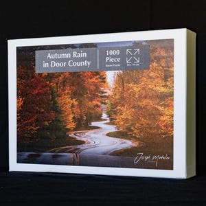 May include: A rectangular jigsaw puzzle box featuring a 1000 piece count and the title Autumn Rain in Door County. The box art displays a winding road through a forest with vibrant orange and yellow autumn foliage. Dimensions are 50 x 70 cm.