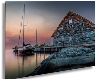May include: A sailboat is docked at a wooden pier with a colorful building in the background. The building is covered in graffiti and has a string of lights across the front. The sky is a vibrant orange and pink.