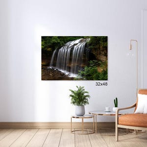 May include: A photograph of a waterfall cascading over rocks in a lush green forest. The water is white and foamy, and the rocks are brown and mossy. The forest is dense and green, with tall trees and ferns. The image is taken from a low angle, looking up at the waterfall.