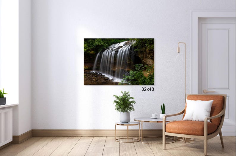 May include: A photograph of a waterfall cascading over rocks in a lush green forest. The water is white and foamy, and the rocks are brown and mossy. The forest is dense and green, with tall trees and ferns. The image is taken from a low angle, looking up at the waterfall.