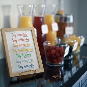 May include: A framed print with the Spanish words "Soy amable", "Soy valiente", "Soy honesto", "Soy inteligente", "Soy fuerte", "Soy creativo", and "Soy amado" with English translations below each phrase. The print is on a table with a bowl of raspberries and a bowl of blueberries.