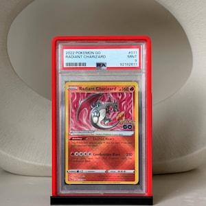 May include: A graded 2022 Pok&eacute;mon GO Radiant Charizard card in a red case. The card features a fiery background and a depiction of Charizard. The card is graded as Mint 9 by PSA, with the number 92192611.