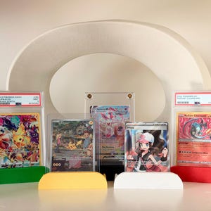 May include: Several graded Pokémon trading cards in protective cases, displayed on colorful stands. The cards feature various characters and designs, including Pikachu and Charizard. The cases are supported by green, yellow, white, and red stands.